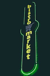 A vertical neon sign with the words 'bistro market' displayed in bright yellow against a dark background. The outline of a shopping cart is integrated into the design above the text, and the sign is surrounded by a glowing green border.