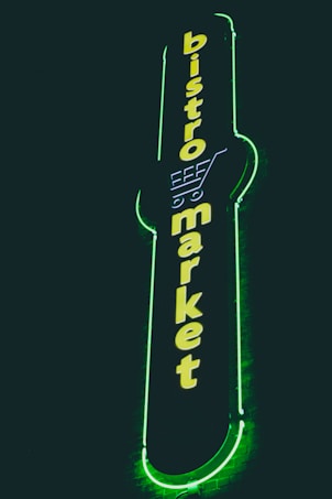 A vertical neon sign with the words 'bistro market' displayed in bright yellow against a dark background. The outline of a shopping cart is integrated into the design above the text, and the sign is surrounded by a glowing green border.