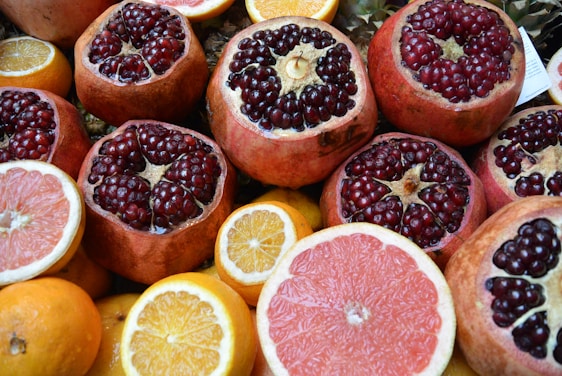 An assortment of fresh fruits featuring prominently cut pomegranates with ruby red seeds glistening, surrounded by slices and whole citrus fruits like oranges and grapefruits. The vibrant colors of the fruits create an appealing and fresh presentation.