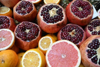 An assortment of fresh fruits featuring prominently cut pomegranates with ruby red seeds glistening, surrounded by slices and whole citrus fruits like oranges and grapefruits. The vibrant colors of the fruits create an appealing and fresh presentation.