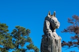 A wooden sculpture of an animal with detailed carvings, set against a clear blue sky. Trees with green and brown foliage are visible in the background.