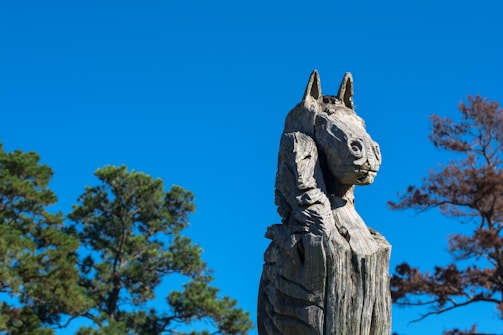 A wooden sculpture of an animal with detailed carvings, set against a clear blue sky. Trees with green and brown foliage are visible in the background.