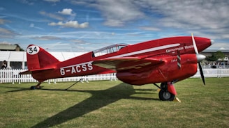 A red vintage aircraft with the registration number G-ACSS is displayed on a grassy field. The plane has the number 34 painted on its tail and the name 'Grosvenor House' is visible on its fuselage. White picket fences and tents are set up in the background under a partly cloudy sky.