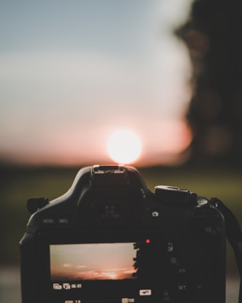 A digital camera captures a vivid sunset. The scene is reflected on the camera screen, emphasizing the vibrant colors of the setting sun against a serene landscape. The silhouette of the camera is prominently featured, with the sun positioned at its center, creating a balanced composition.