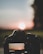 A digital camera captures a vivid sunset. The scene is reflected on the camera screen, emphasizing the vibrant colors of the setting sun against a serene landscape. The silhouette of the camera is prominently featured, with the sun positioned at its center, creating a balanced composition.