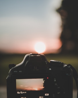 A digital camera captures a vivid sunset. The scene is reflected on the camera screen, emphasizing the vibrant colors of the setting sun against a serene landscape. The silhouette of the camera is prominently featured, with the sun positioned at its center, creating a balanced composition.
