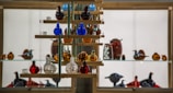 A glass display case filled with colorful glass figurines arranged on wooden shelves. The figurines resemble birds and other animals, predominantly in shades of blue, red, amber, and clear glass. The background is illuminated, highlighting the vibrant colors and intricate details of the glass art pieces.