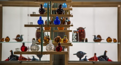 A glass display case filled with colorful glass figurines arranged on wooden shelves. The figurines resemble birds and other animals, predominantly in shades of blue, red, amber, and clear glass. The background is illuminated, highlighting the vibrant colors and intricate details of the glass art pieces.