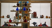 A glass display case filled with colorful glass figurines arranged on wooden shelves. The figurines resemble birds and other animals, predominantly in shades of blue, red, amber, and clear glass. The background is illuminated, highlighting the vibrant colors and intricate details of the glass art pieces.