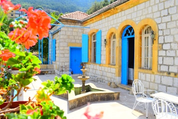 A charming stone house with bright blue doors and arched windows, surrounded by vibrant flowers in the foreground. A small fountain is situated on the patio, which has white metal chairs and tables. The backdrop features green hills and a clear sky.