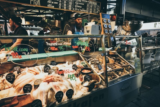 A bustling seafood market stall with a variety of fresh fish and shellfish displayed on ice. Chalkboard menus above the counter list various seafood items and prices. Two workers, wearing aprons and hats, are behind the counter attending to customers. The counter contains oysters, fish fillets, and other seafood delicacies, with labels indicating type and price. The ambiance suggests a lively market atmosphere.
