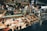 A bustling seafood market stall with a variety of fresh fish and shellfish displayed on ice. Chalkboard menus above the counter list various seafood items and prices. Two workers, wearing aprons and hats, are behind the counter attending to customers. The counter contains oysters, fish fillets, and other seafood delicacies, with labels indicating type and price. The ambiance suggests a lively market atmosphere.