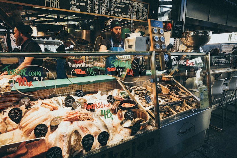 A bustling seafood market stall with a variety of fresh fish and shellfish displayed on ice. Chalkboard menus above the counter list various seafood items and prices. Two workers, wearing aprons and hats, are behind the counter attending to customers. The counter contains oysters, fish fillets, and other seafood delicacies, with labels indicating type and price. The ambiance suggests a lively market atmosphere.