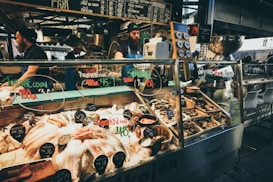 A bustling seafood market stall with a variety of fresh fish and shellfish displayed on ice. Chalkboard menus above the counter list various seafood items and prices. Two workers, wearing aprons and hats, are behind the counter attending to customers. The counter contains oysters, fish fillets, and other seafood delicacies, with labels indicating type and price. The ambiance suggests a lively market atmosphere.
