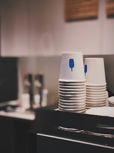 A stack of white paper cups with blue designs sits on a countertop in a kitchen or cafe environment. The background is softly blurred, featuring additional kitchen items and subtle lighting adding a cozy atmosphere.