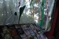 A weathered window with broken glass segments, some of which are stained with colorful paint splatters in green, orange, and blue shades. There are multiple labels of beer or alcohol bottles stuck on one of the surfaces near the bottom, each displaying intricate designs and various brands.