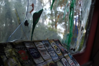 A weathered window with broken glass segments, some of which are stained with colorful paint splatters in green, orange, and blue shades. There are multiple labels of beer or alcohol bottles stuck on one of the surfaces near the bottom, each displaying intricate designs and various brands.