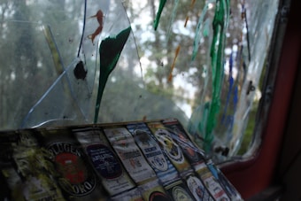 A weathered window with broken glass segments, some of which are stained with colorful paint splatters in green, orange, and blue shades. There are multiple labels of beer or alcohol bottles stuck on one of the surfaces near the bottom, each displaying intricate designs and various brands.