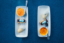 Two white rectangular plates are placed on a textured blue surface, each with a small silver spoon resting on top. Each plate features a creme brulee with a golden-brown caramelized top, a rectangular chocolate dessert with a blue and white cream garnish, and a madeleine. There is a sprinkling of granulated sugar on one of the madeleines.
