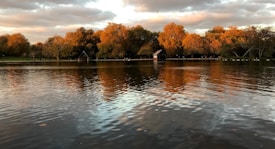 A serene lake with calm waters reflecting the vibrant autumn foliage of trees surrounding it. The sky is overcast with soft, fluffy clouds capturing the warm hues of sunset. A small wooden structure is visible near the water's edge, adding a rustic touch to the landscape.