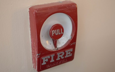 A red fire alarm pull station mounted on a beige wall, featuring a noticeable circle with the word 'PULL' in white letters and the word 'FIRE' at the bottom. The surface shows signs of wear.