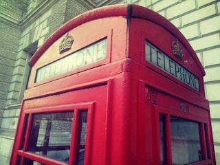 A classic red British telephone booth with the word 'TELEPHONE' displayed above the windows. The booth is positioned against a stone building facade featuring intricate patterns.