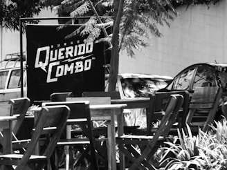 A black and white photograph featuring a small outdoor seating area with wooden tables and chairs. In the background, there is a large sign that reads 'Burger Querido Combo.' Several cars are parked near the sign, and trees with leafy branches create shade over the area.
