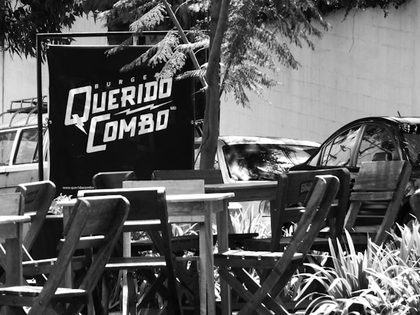 A black and white photograph featuring a small outdoor seating area with wooden tables and chairs. In the background, there is a large sign that reads 'Burger Querido Combo.' Several cars are parked near the sign, and trees with leafy branches create shade over the area.