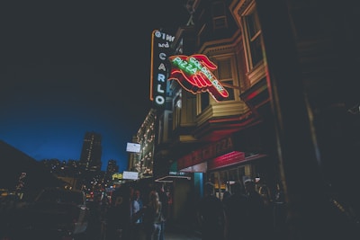 Neon signs glow outside an urban establishment at night. The signs are attached to a building with a distinct architectural style. People are gathered on the sidewalk beneath the signs, creating an atmosphere of nightlife and social gathering.