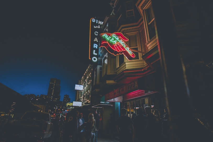 Neon signs glow outside an urban establishment at night. The signs are attached to a building with a distinct architectural style. People are gathered on the sidewalk beneath the signs, creating an atmosphere of nightlife and social gathering.