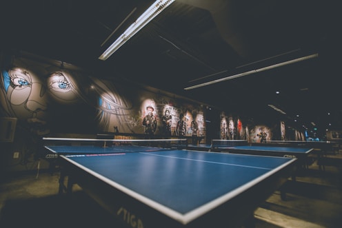 A dimly lit room with two blue ping pong tables dominates the foreground. The walls are adorned with murals and graffiti, featuring artistic portraits and abstract designs. The ceiling has modern light fixtures that cast an industrial ambiance over the space.