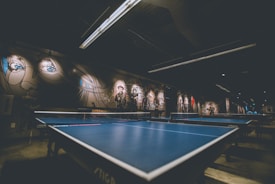 A dimly lit room with two blue ping pong tables dominates the foreground. The walls are adorned with murals and graffiti, featuring artistic portraits and abstract designs. The ceiling has modern light fixtures that cast an industrial ambiance over the space.