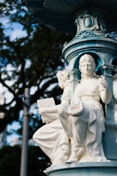 A detailed sculpture of a seated figure adorned in classical attire is part of a larger ornamented fountain structure. The sculpture appears to be made of marble or another white stone. The background includes blurred foliage of trees, and the image is captured with natural daylight, highlighting the intricate carvings and designs.