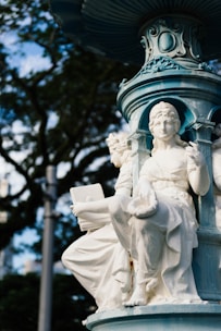 A detailed sculpture of a seated figure adorned in classical attire is part of a larger ornamented fountain structure. The sculpture appears to be made of marble or another white stone. The background includes blurred foliage of trees, and the image is captured with natural daylight, highlighting the intricate carvings and designs.