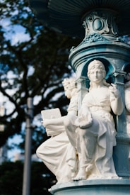 A detailed sculpture of a seated figure adorned in classical attire is part of a larger ornamented fountain structure. The sculpture appears to be made of marble or another white stone. The background includes blurred foliage of trees, and the image is captured with natural daylight, highlighting the intricate carvings and designs.