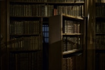 A dimly lit library interior featuring tall wooden bookshelves filled with numerous aged, leather-bound books. One of the shelves appears to be a hidden door, slightly ajar, revealing a shadowy passageway or hidden room. The ambiance is reminiscent of a classic or historical setting, suggesting mystery and intrigue.