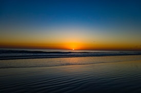 A serene beach scene at sunset with a vibrant gradient sky transitioning from deep blue to warm orange. Gentle waves reach the shore, and several birds fly across the horizon, creating a peaceful atmosphere.