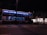 A large double-decker bus with illuminated blue interior lights is stationary on a city street at night. The bus is advertising a movie or event named 'Midnight Special.' It is surrounded by urban buildings, including a store with bright windows. Traffic lights are visible, and the scene is relatively quiet with minimal movement.