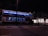 A large double-decker bus with illuminated blue interior lights is stationary on a city street at night. The bus is advertising a movie or event named 'Midnight Special.' It is surrounded by urban buildings, including a store with bright windows. Traffic lights are visible, and the scene is relatively quiet with minimal movement.