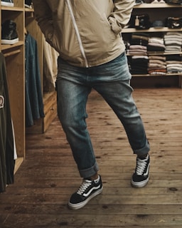 A person wearing a beige jacket and blue jeans is standing casually with hands in pockets. They are in a clothing store, evident from the neatly stacked clothes on shelves in the background. The floor is wooden, adding a warm and rustic feel.