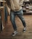 A person wearing a beige jacket and blue jeans is standing casually with hands in pockets. They are in a clothing store, evident from the neatly stacked clothes on shelves in the background. The floor is wooden, adding a warm and rustic feel.