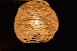 A spherical lamp made of intricately woven material that surrounds a bulb, casting a warm, inviting glow against a dark background.