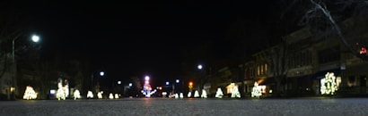 A nighttime street scene lined with Christmas lights and decorations. Small illuminated trees are placed along the sidewalk, and buildings are subtly lit in the background. Streetlights cast a gentle glow over the scene, creating a festive and serene atmosphere.