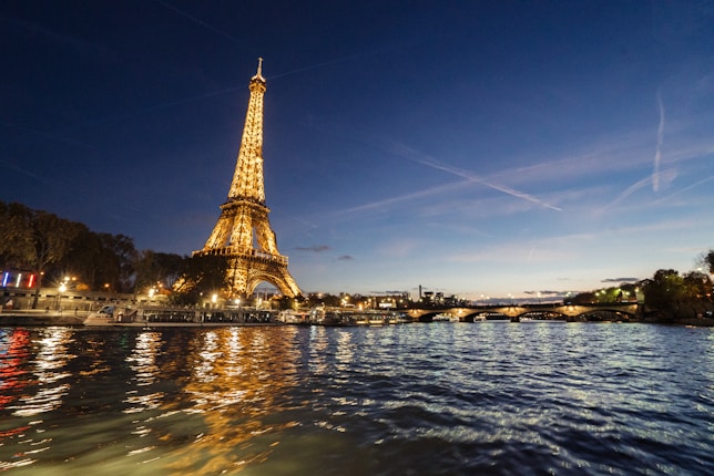 The Eiffel Tower is illuminated at night, standing tall against a deep blue sky. The Seine River in the foreground reflects the lights from the tower and nearby cityscape. A bridge can be seen to the right, along with silhouettes of trees and buildings.