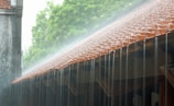 A sloping roof with red tiles experiences heavy rain, creating cascading streams of water flowing down across the eaves. In the background, lush green trees are faintly visible through a misty atmosphere.