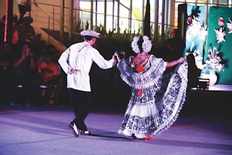 A man and a woman are performing a traditional dance together in an outdoor setting. The woman is wearing a colorful, intricately patterned dress with floral designs and a headdress adorned with flowers, while the man is dressed in a white shirt, dark pants, and a hat. The background features an audience seated in the dark and a decorated wall with plants and flower motifs.