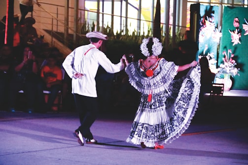 A man and a woman are performing a traditional dance together in an outdoor setting. The woman is wearing a colorful, intricately patterned dress with floral designs and a headdress adorned with flowers, while the man is dressed in a white shirt, dark pants, and a hat. The background features an audience seated in the dark and a decorated wall with plants and flower motifs.