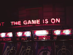 Neon lights display the phrase 'THE GAME IS ON' above a series of arcade machines. These machines are labeled 'ICE BALL' and display scores and numbers in bright colors. The setting appears to be a dimly lit arcade, creating an energetic and exciting atmosphere.