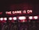 Neon lights display the phrase 'THE GAME IS ON' above a series of arcade machines. These machines are labeled 'ICE BALL' and display scores and numbers in bright colors. The setting appears to be a dimly lit arcade, creating an energetic and exciting atmosphere.