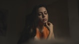 A woman with long dark hair is sitting with her eyes closed, appearing deep in thought or contemplation. She wears an orange top, and her hand is gently touching her chin. The lighting is dim, casting a soft, moody shadow around her.